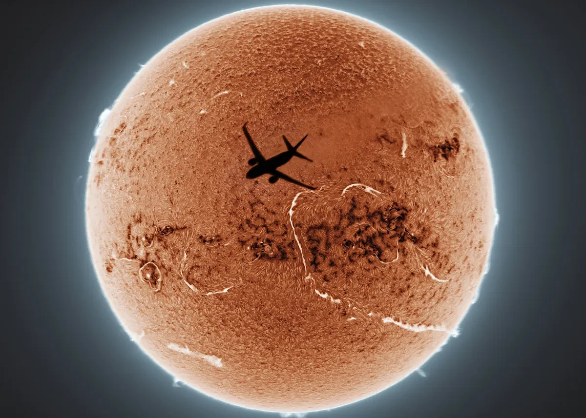 Photo of the Sun in bright orange outlined in white, with a plane silhouette appearing to be heading into the Sun